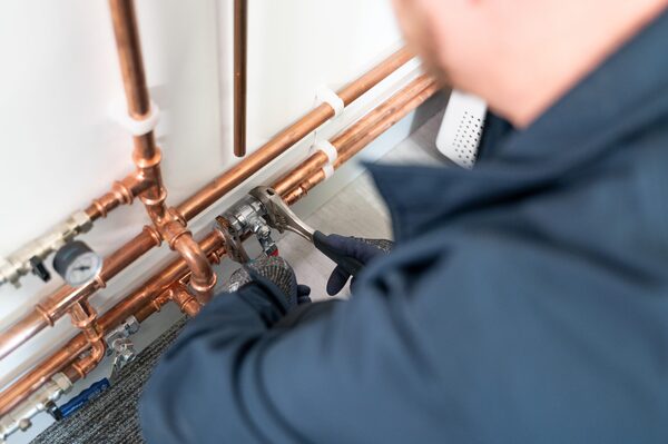 A heating engineer in a navy jacket using a spanner on copper pipework connected to a boiler
