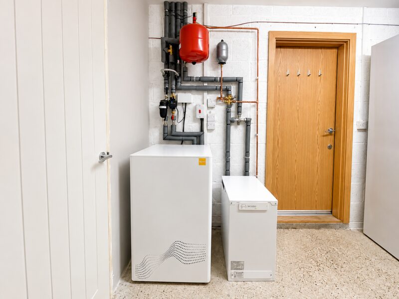 A white ZEB heat battery boiler installed in a utility room with grey pipework, expansion vessels and a wooden door