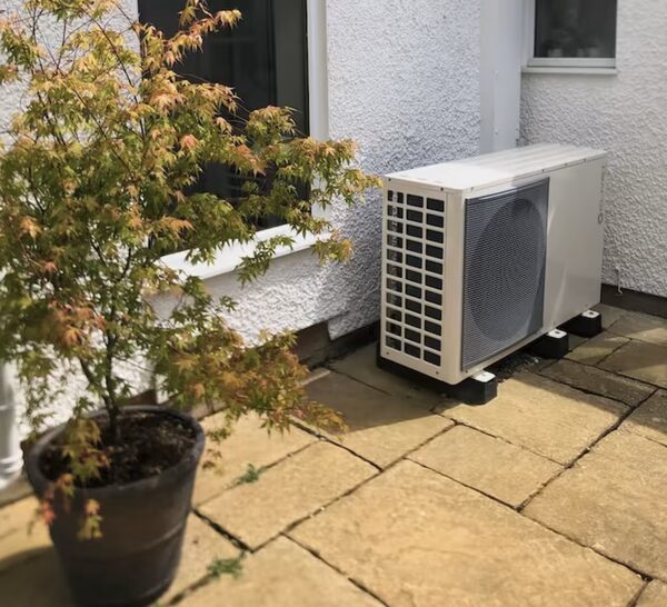 An air source heat pump unit installed outside a house on a paved patio next to a potted acer tree