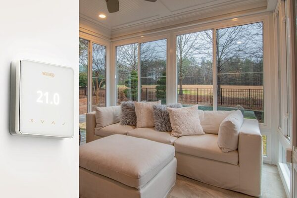 A Warmup wall thermostat displaying 21.0 mounted on a wall beside a bright conservatory-style living room with a cream corner sofa