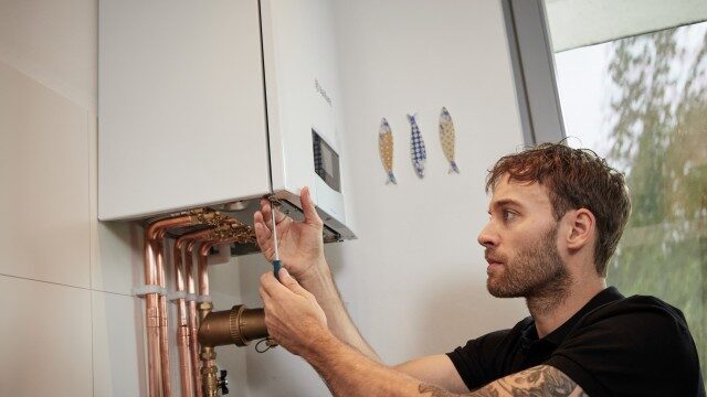 An engineer using a screwdriver to service a white wall-mounted Vaillant combi boiler with copper pipework in a home
