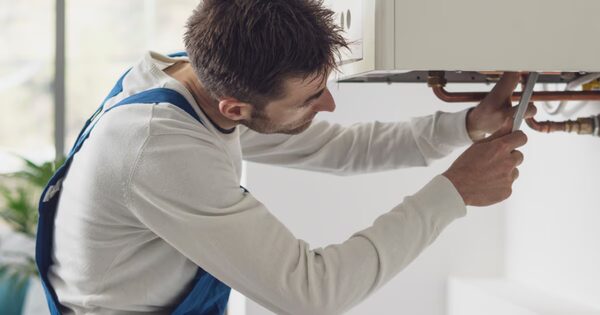 A heating engineer in blue overalls using a spanner to work on the copper pipework beneath a wall-mounted gas boiler