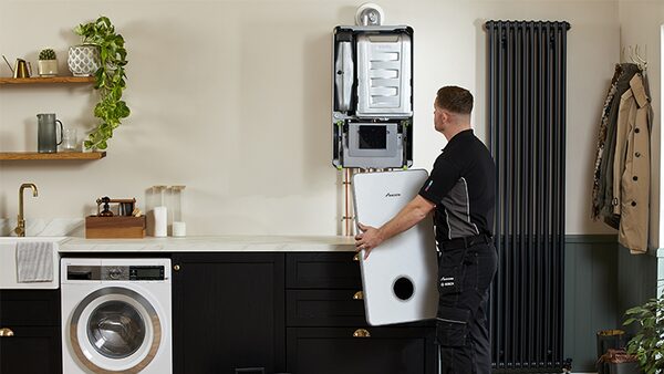 An engineer fitting a white Worcester Bosch system boiler on a kitchen wall, holding the front casing panel