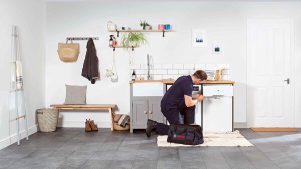 A technician kneeling in a utility room servicing a white boiler under a worktop, with a Bosch tool bag beside him