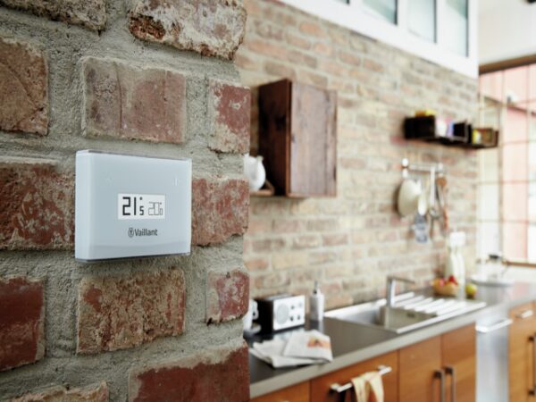 A white Vaillant vSMART smart thermostat mounted on an exposed brick wall showing 21.5°C, with a kitchen in the background