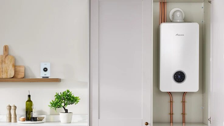 A white Worcester Bosch combi boiler installed in a kitchen cupboard with copper pipework, next to a kitchen counter with a plant and a wall-mounted thermostat