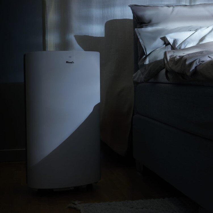 A white Woods portable air conditioner standing on the floor next to a bed in a dimly lit bedroom