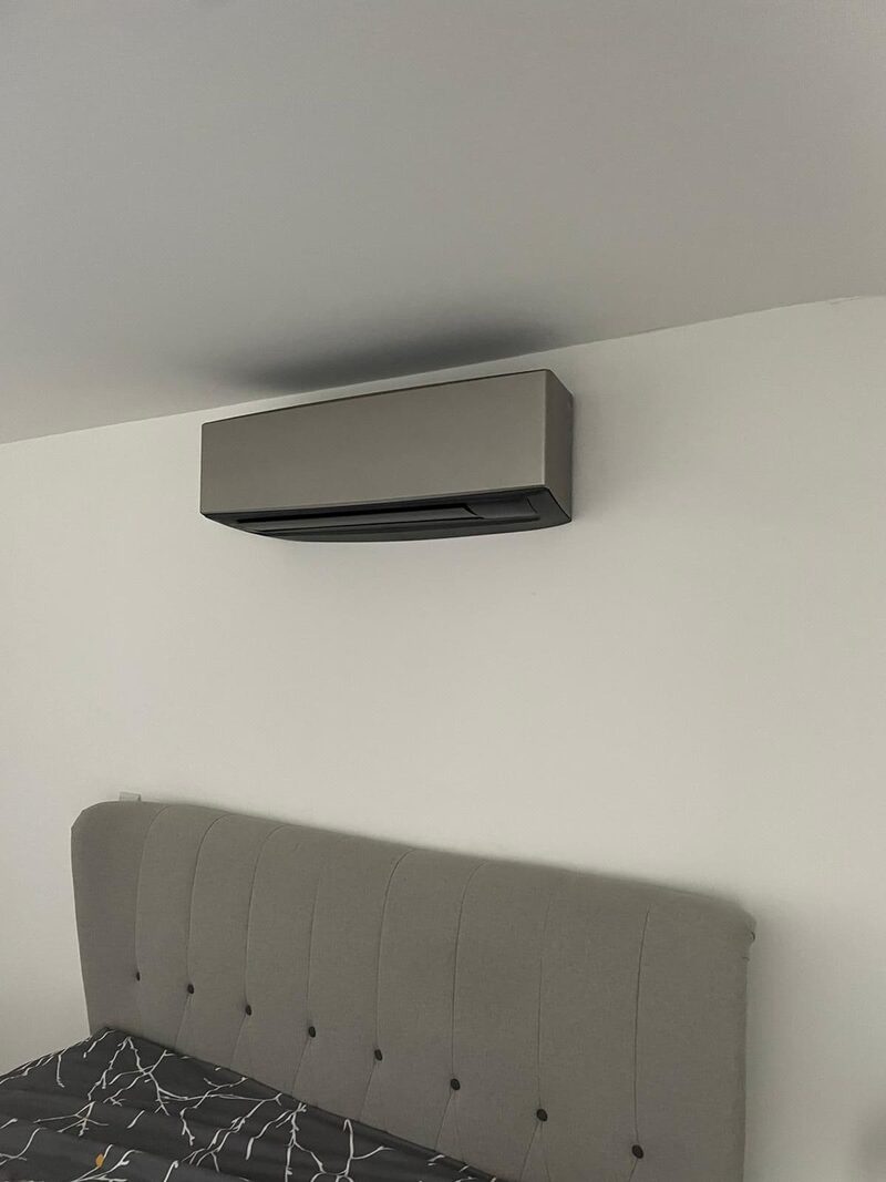A wall-mounted air conditioning / heat pump indoor unit installed above a bed in a bedroom