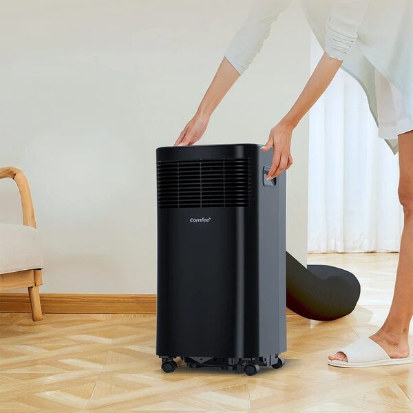 A woman positioning a black Comfee portable air conditioner with exhaust hose in a home room