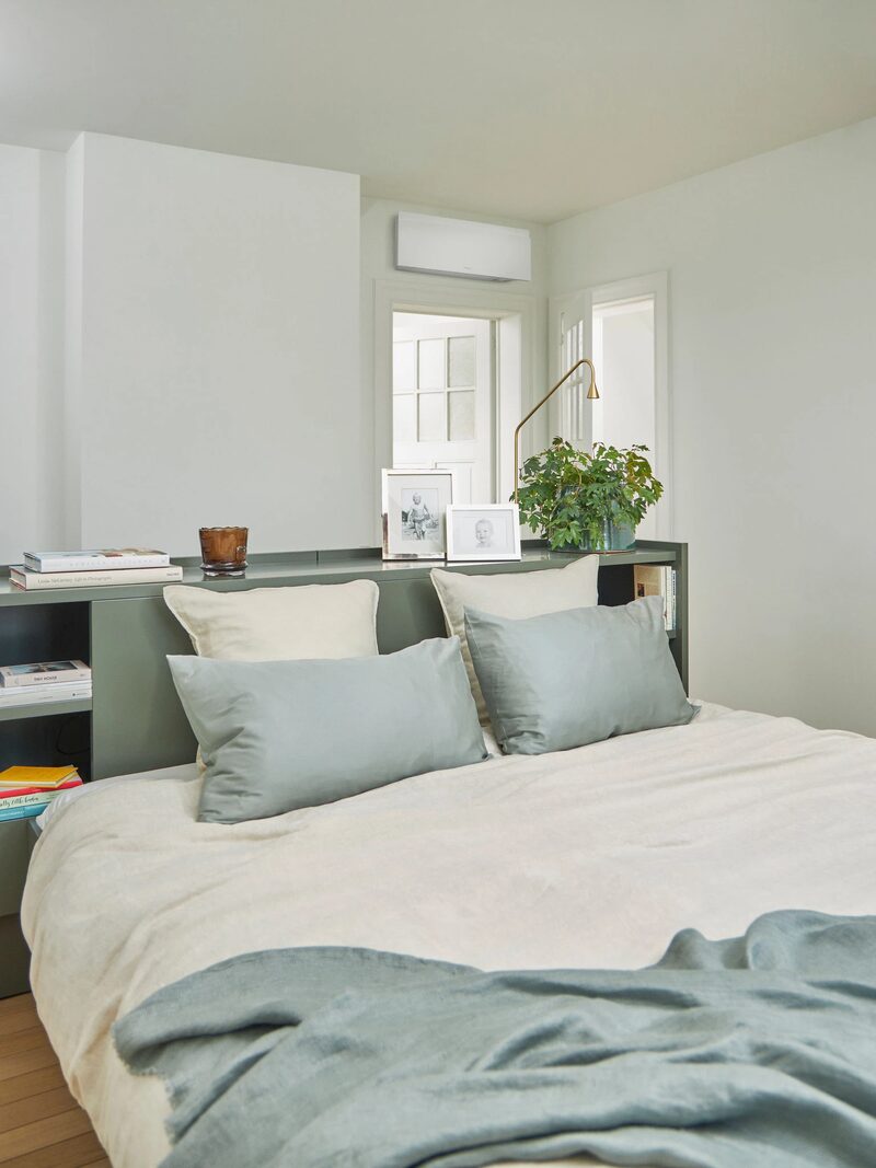 A white wall-mounted air conditioning unit installed high on the wall above a bed in a bright bedroom