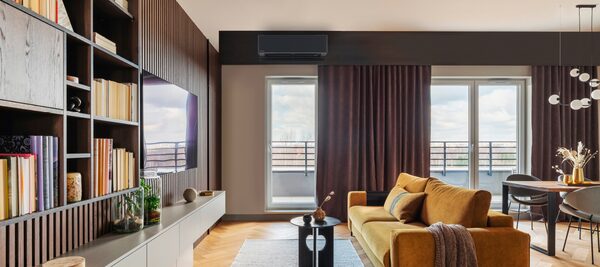 A wall-mounted air conditioning unit installed high on the wall of a modern living room with a mustard sofa and bookshelves
