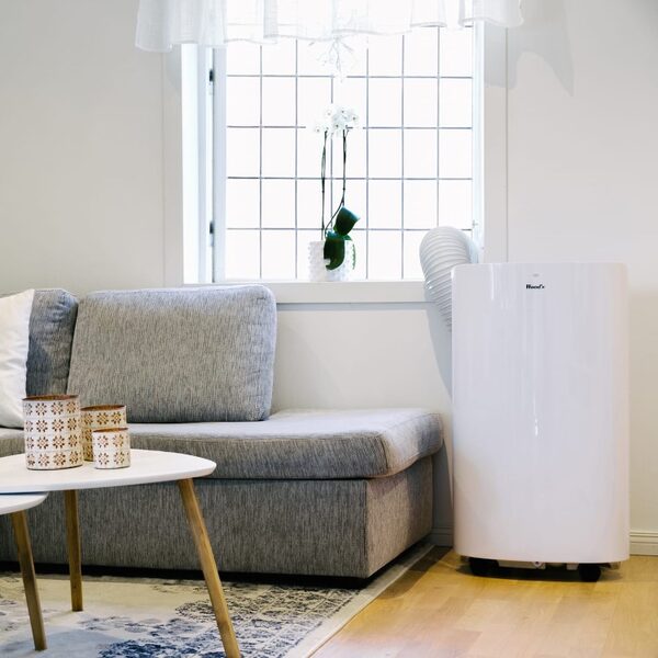 A white Woods portable air conditioner standing in a bright living room next to a grey sofa, with its exhaust hose running to the window