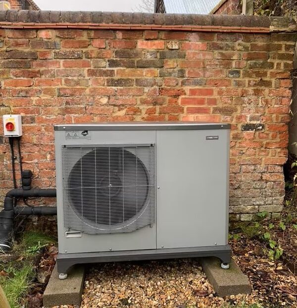 A grey air source heat pump unit installed outside against a red brick wall with pipework and an isolation switch