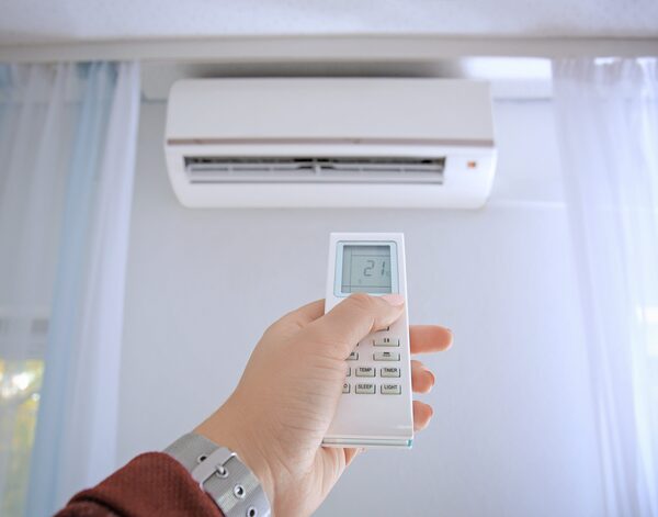 A hand holding a remote control pointing at a wall-mounted air conditioning unit above a window with sheer curtains