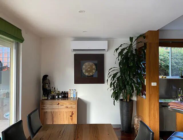 A white wall-mounted split air conditioning unit installed high on the wall of a dining room with a wooden table, plant and cabinet