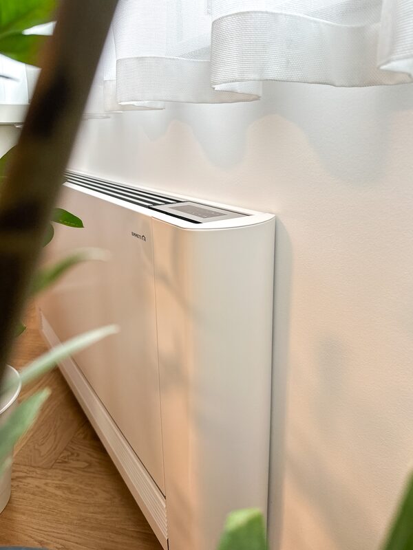 A white Kronoterm indoor heat pump fan coil unit installed against a wall in a room, partially obscured by a houseplant