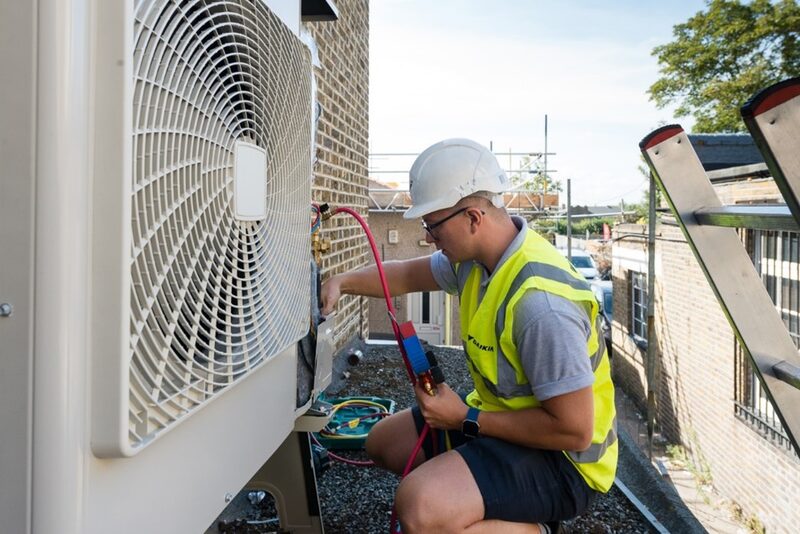 An engineer in a hi-vis vest and hard hat checks a Daikin air source heat pump outdoor unit with gauges on a rooftop