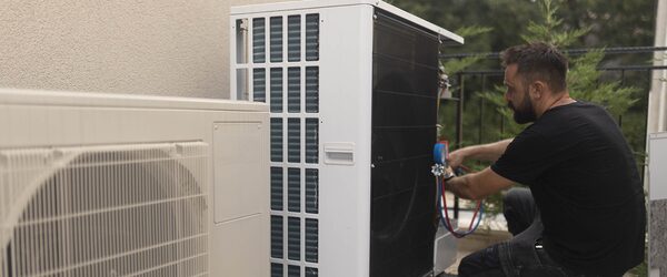 A technician working on an outdoor air conditioning / heat pump unit with gauges, next to another unit on a balcony