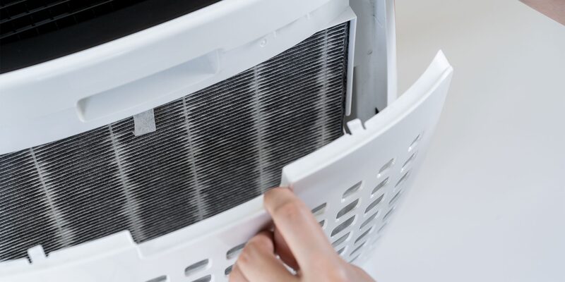 A hand removing the front panel of a portable air conditioner to reveal a dusty grey filter