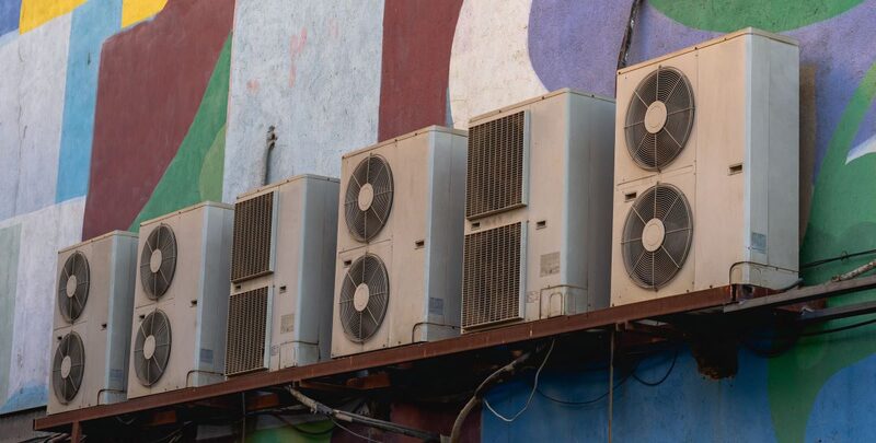 A row of outdoor air conditioning condenser units mounted on a rack against a colourfully painted wall