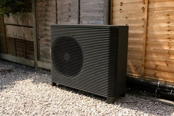 A dark grey outdoor air source heat pump unit with a circular fan grille standing on gravel beside a wooden fence