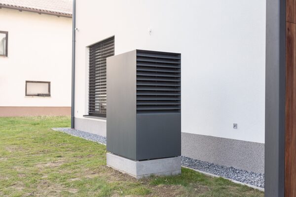 A grey outdoor air source heat pump unit installed on a concrete plinth beside the exterior wall of a house
