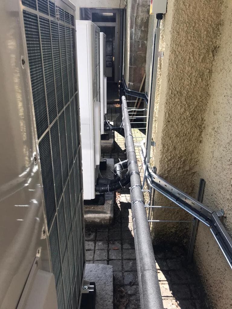 Two air source heat pump units installed in a narrow side passage beside a house wall with insulated pipework