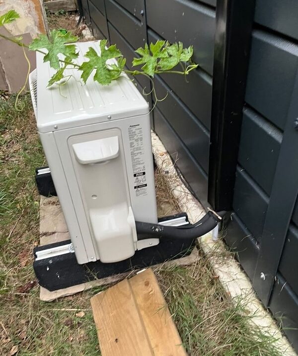 A Panasonic air conditioning heat pump outdoor unit installed on a base beside a dark-clad building wall