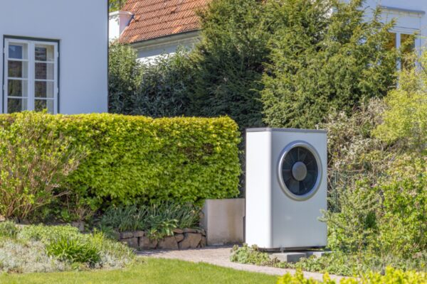 A white air source heat pump unit installed in a garden beside a house with hedges and trees