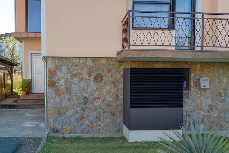 A dark grey heat pump outdoor unit installed against the stone base of a house exterior wall