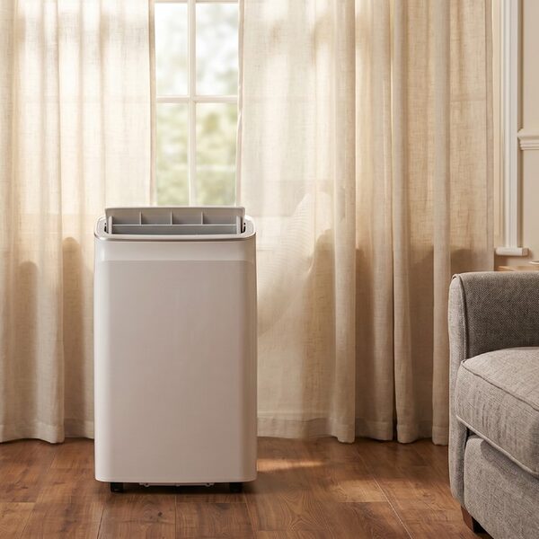 A white electriQ portable air conditioner standing on a wooden floor in a living room beside a sofa and curtained window
