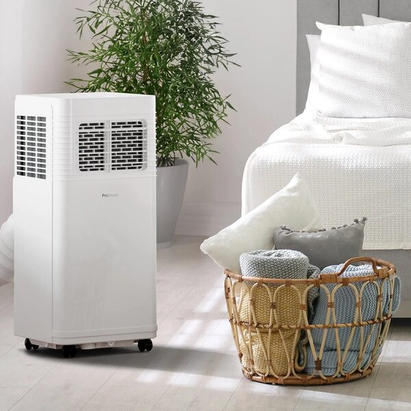 A white Pro Breeze portable air conditioner standing in a bright bedroom next to a bed, plant and basket of blankets