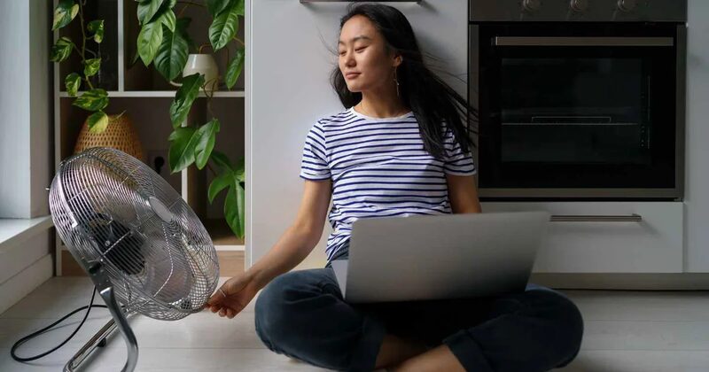 A woman sitting on a kitchen floor with a laptop, cooling off in front of an electric floor fan