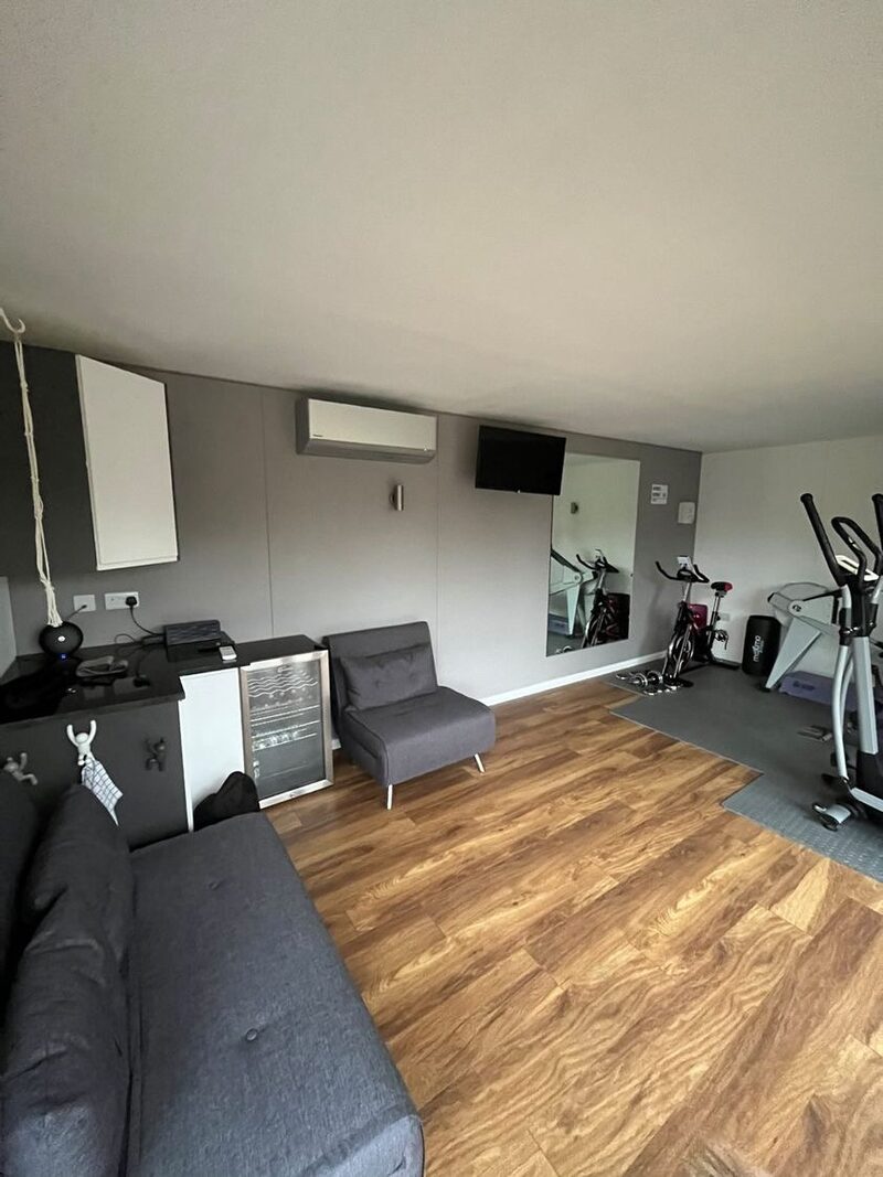 A wall-mounted air conditioning unit installed high on the wall of a home gym and living room with exercise bikes, a sofa and wood flooring