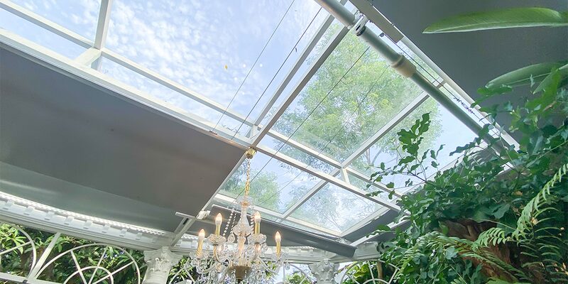Roof blinds partly drawn across the glass roof of a conservatory with a chandelier and plants
