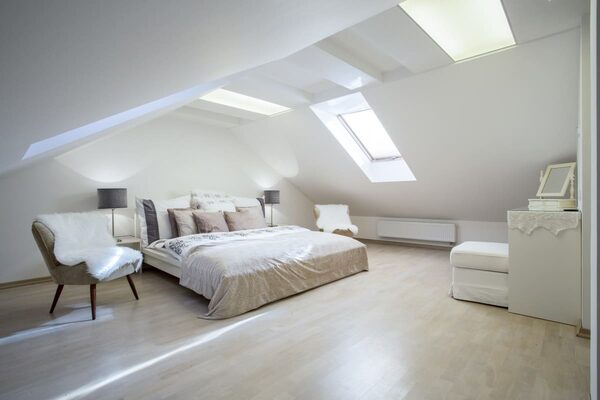 A bright loft-conversion bedroom with pale wooden flooring, a bed, chairs and a wall radiator under a sloped ceiling with roof windows