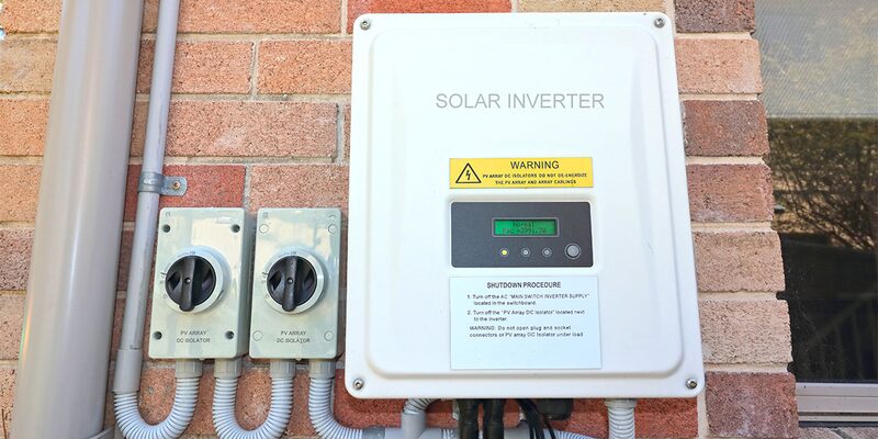 A white solar inverter labelled 'SOLAR INVERTER' with warning labels and a display, mounted on a brick wall next to two PV array DC isolator switches