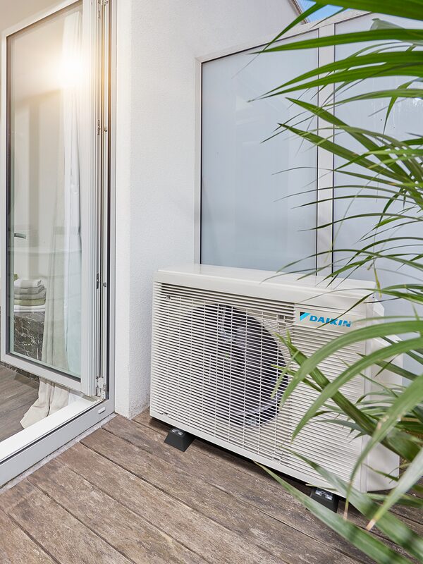 A white Daikin air source heat pump outdoor unit installed on a wooden terrace beside a house wall