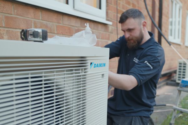 An engineer in a Daikin polo shirt installing an air source heat pump outdoor unit outside a brick house