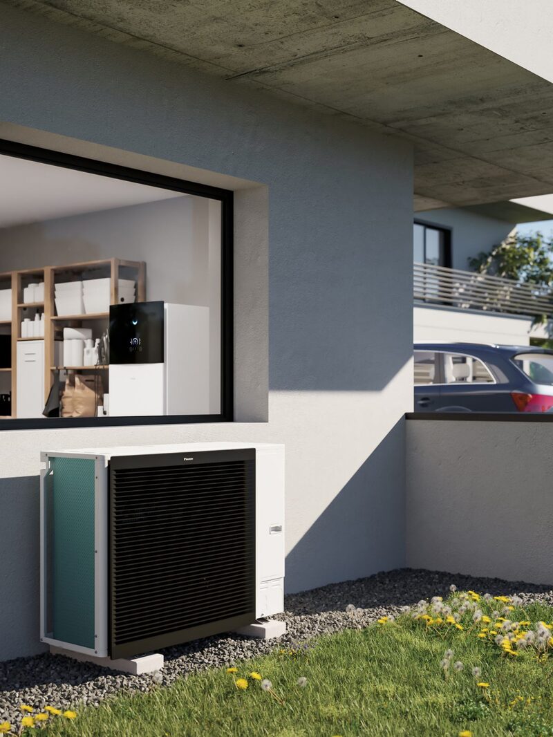 A Daikin air-to-water heat pump unit installed outside a modern house, with an indoor unit visible through the window