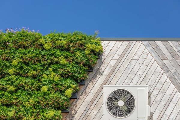A white air source heat pump unit mounted on a weathered timber-clad wall beside a green planted wall under a blue sky