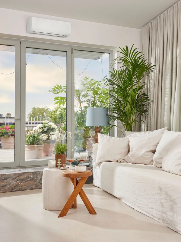 A white wall-mounted air conditioning unit installed high on the wall of a bright living room with a sofa and large glass doors