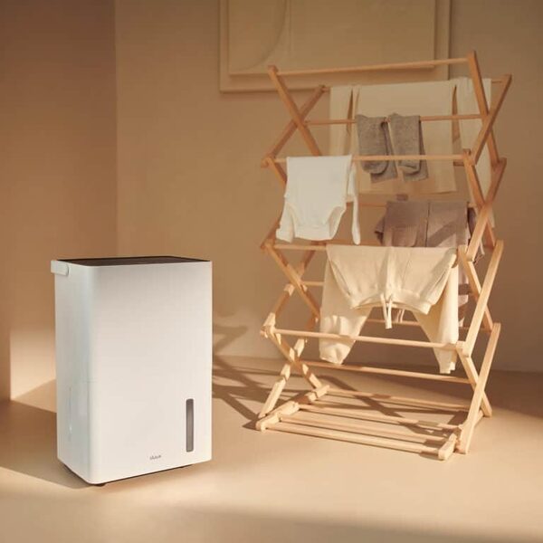 A white Duux Bora dehumidifier standing next to a wooden clothes airer with laundry drying in a warm-toned room