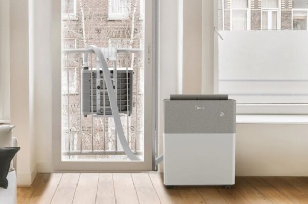 A Midea portable split air conditioner standing in a room with its outdoor unit hung on a balcony railing and the connecting hose running through the window