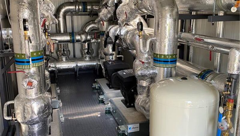 A district heating plant room with insulated pipework, pumps and a white expansion vessel inside an energy centre