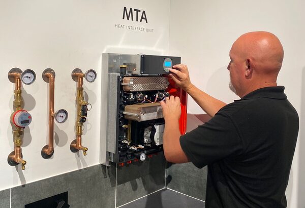 A technician adjusting an MTA heat interface unit mounted on a wall next to copper pipework with pressure gauges and a meter