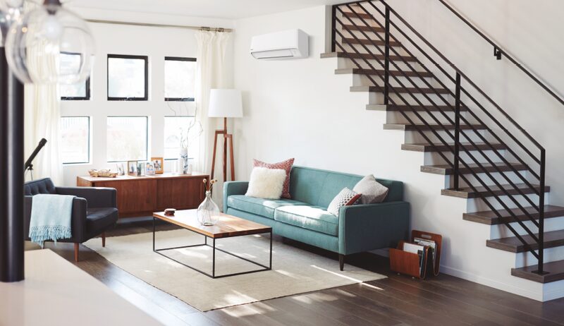 A wall-mounted white air conditioning unit installed high on the wall of a bright modern living room with a teal sofa and staircase