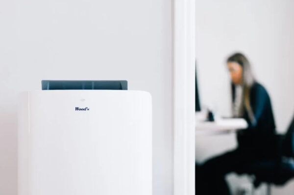 A white Woods portable air conditioner standing in a room with a blurred woman working at a desk in the background