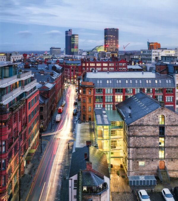 A city skyline at dusk with lit office towers, red brick buildings and traffic light trails on a street