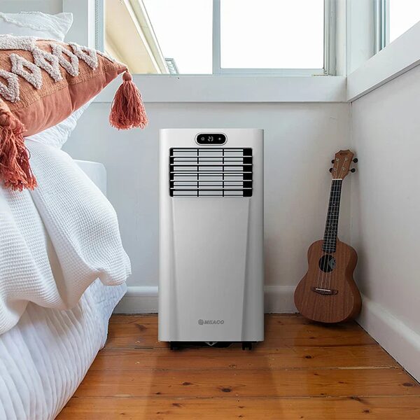 A white Meaco portable air conditioner standing in a bright bedroom next to a bed and a ukulele